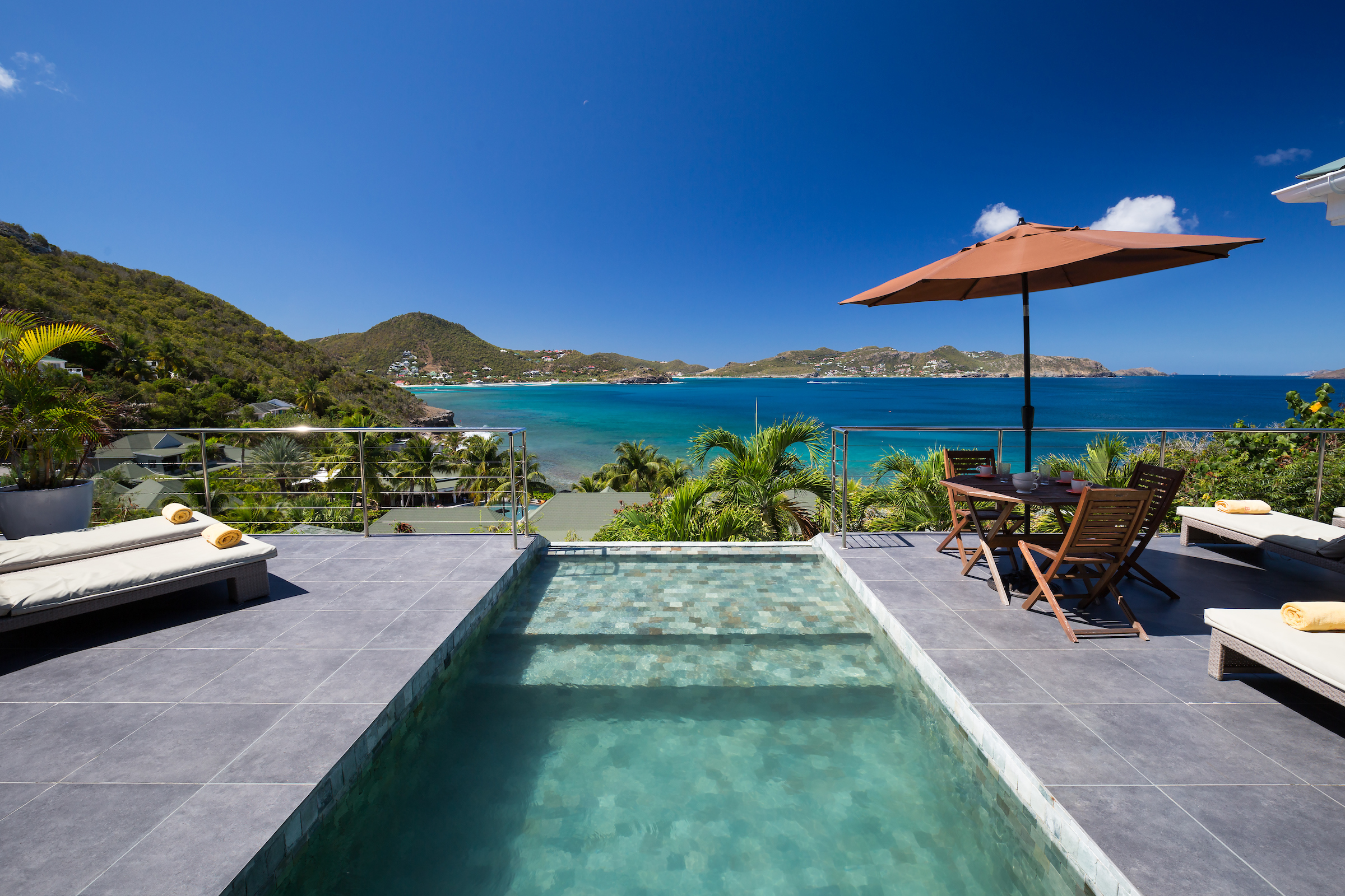 Infinity pool at a St Barts villa overlooking turquoise ocean waters and lush green hills, with sun loungers and shaded dining terrace.