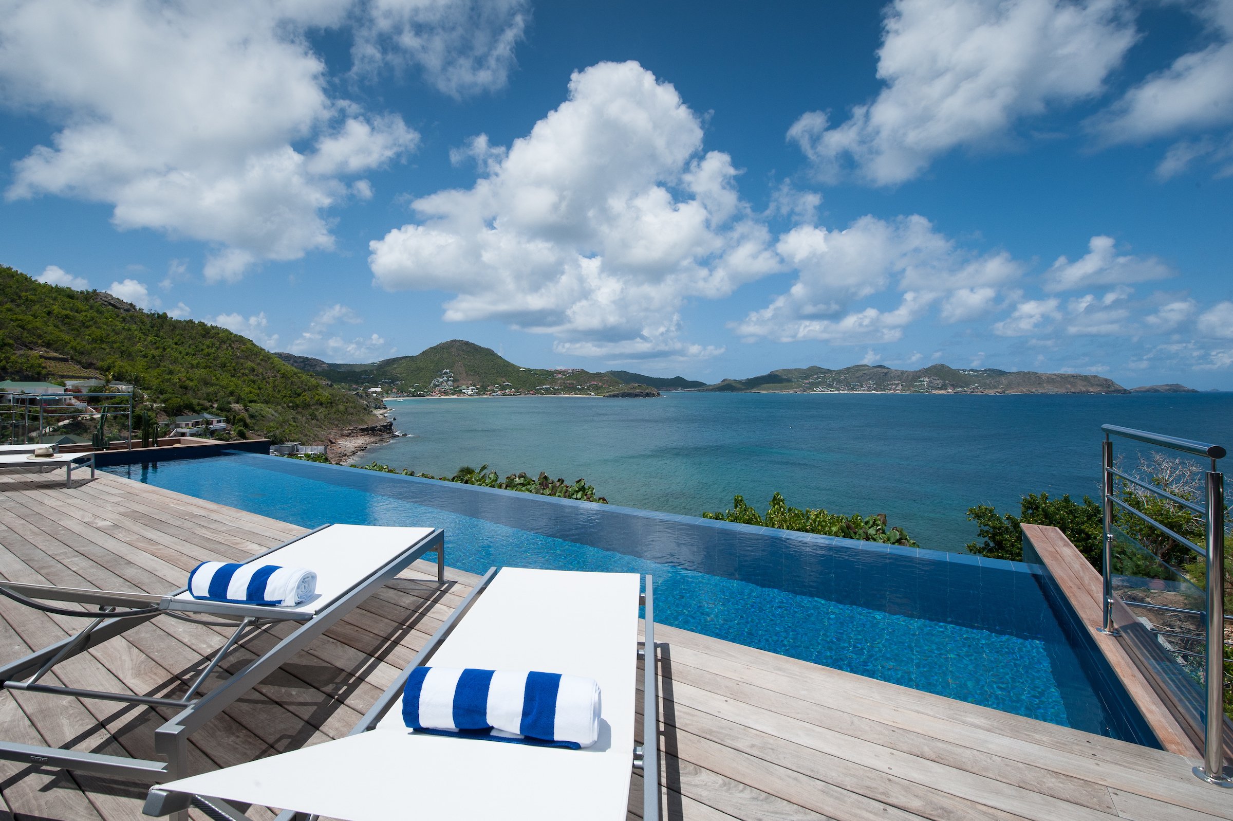 Infinity pool with wooden deck and sun loungers overlooking the Caribbean Sea and rolling green hills in St Barts.