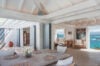 Bright indoor dining space with rustic wooden table, woven placemats, and open doors leading to ocean-view lounge in a St Barts villa.