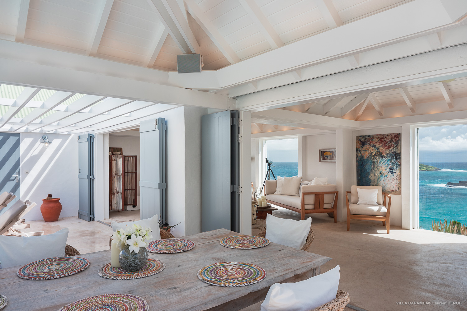 Bright indoor dining space with rustic wooden table, woven placemats, and open doors leading to ocean-view lounge in a St Barts villa.