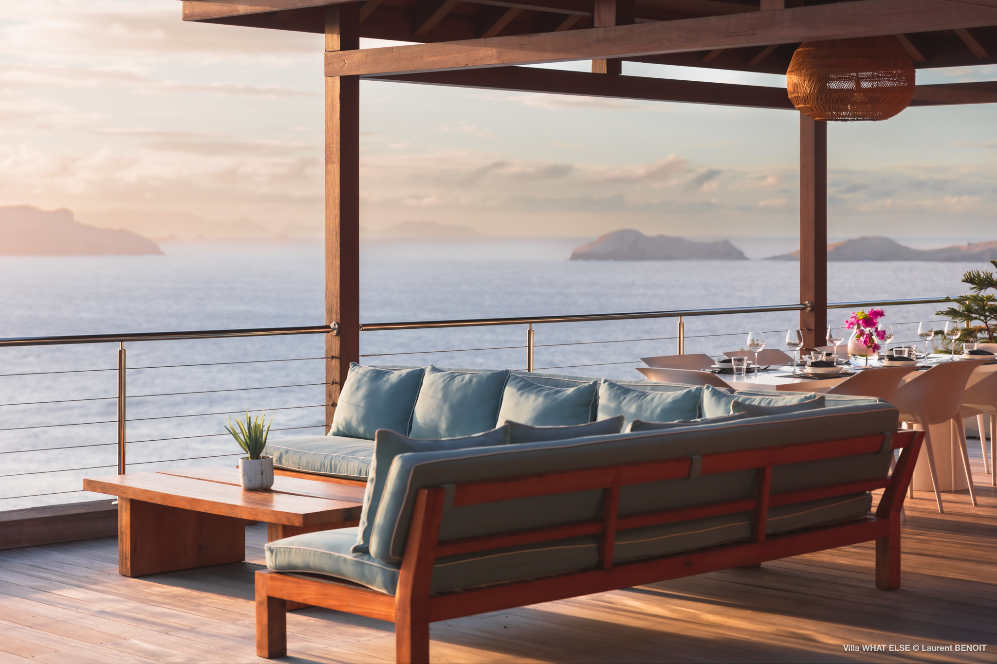 Open-air lounge with teak seating and dining table at Villa What Else overlooking the Caribbean Sea at sunset in St Barts.