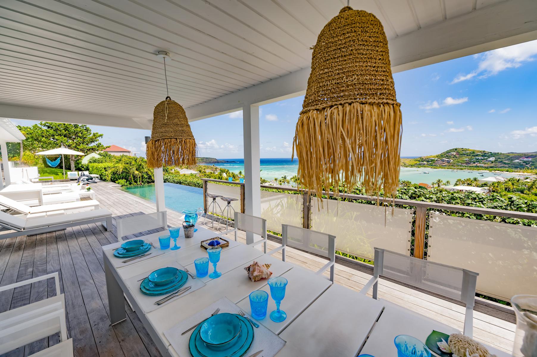Covered outdoor dining terrace in a St Barts villa with turquoise tableware, wicker pendant lights, and panoramic ocean views.