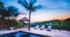 Sunset view over a hillside infinity pool in St Barts, with wooden deck, lounge chairs, and swaying palms against glowing skies.