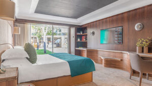 Modern villa room with a large bed, wooden paneling, desk, and glass doors opening to a courtyard.