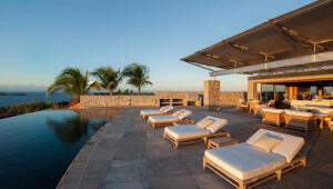 Luxurious villa terrace with sun loungers, infinity pool, palm trees, and ocean view at sunset.