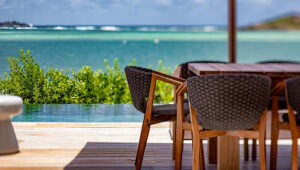 Outdoor wooden dining table with chairs by an infinity pool overlooking turquoise ocean.