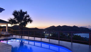 Villa pool terrace overlooking St. Jean Bay at twilight, with mountain silhouettes and city lights.