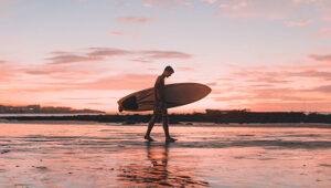 Surfer walking on wet sand carrying a surfboard during a colorful sunset.