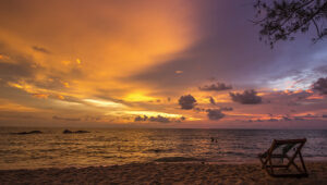 Sunset over the ocean with two people swimming and an empty beach chair on the sandy shore.