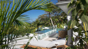 Lush tropical villa patio with a glass railing, cushioned lounge chairs, and a pool surrounded by greenery.