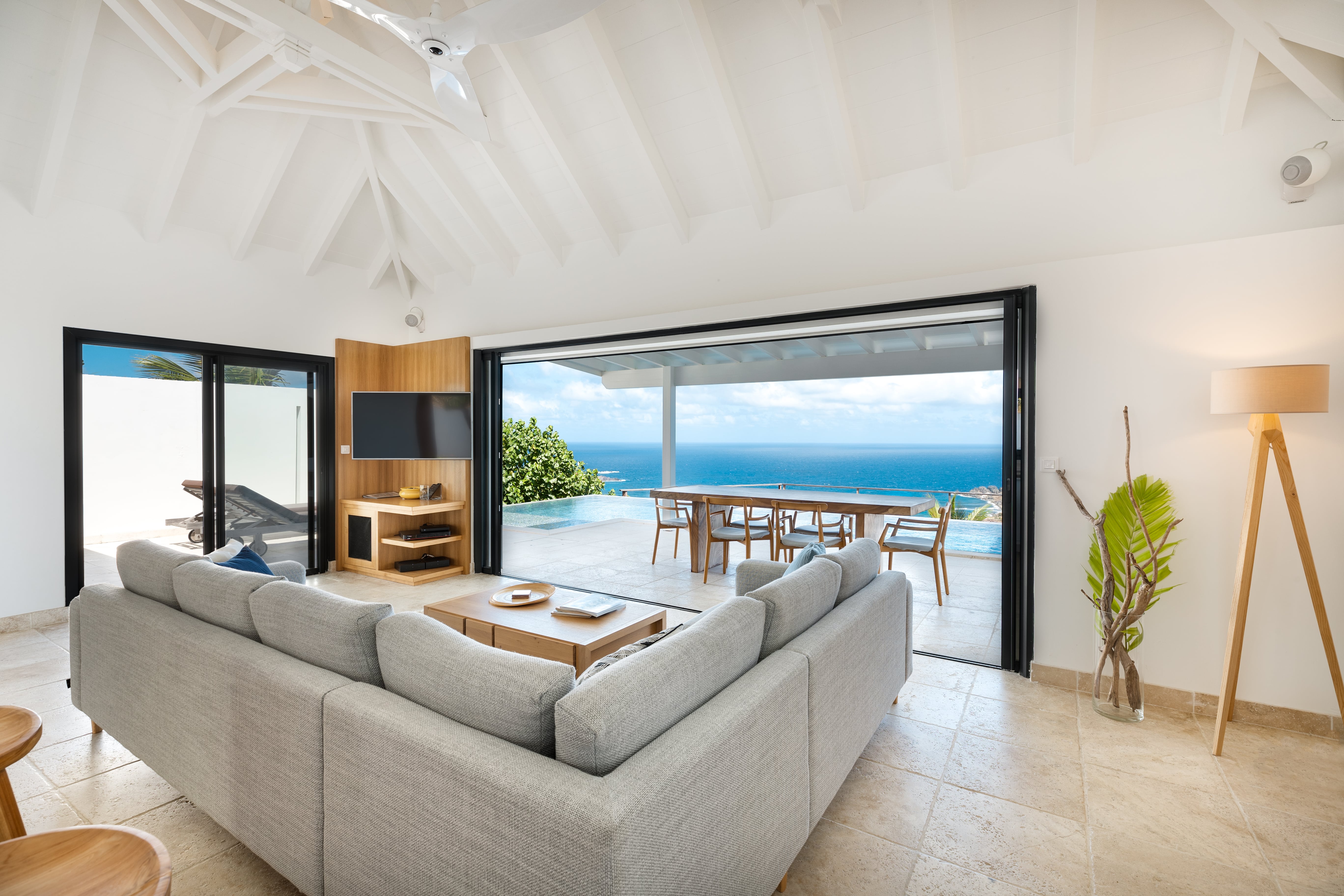 Spacious living room in a St Barts villa with vaulted white ceiling, soft gray sectional sofa, and open view of the infinity pool and Caribbean Sea, perfect for private villa rentals.