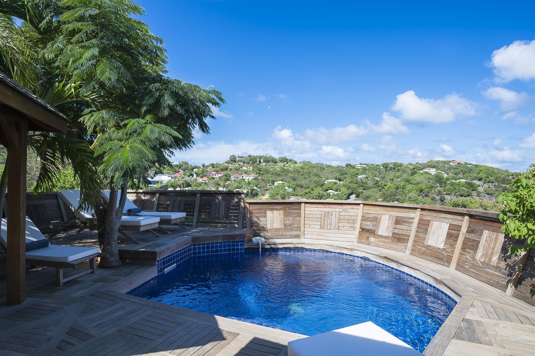 Private villa rentals in St Barts with curved blue pool, wooden deck, and hillside views under clear tropical skies.