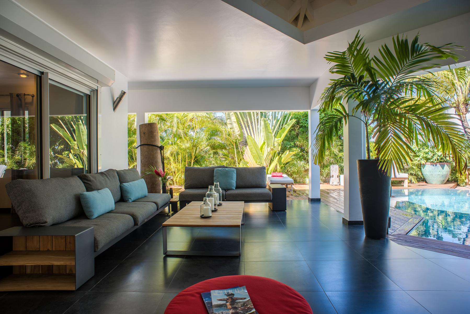 Open-air living area with modern sofas and poolside view surrounded by tropical palms at a St Barts villa rental