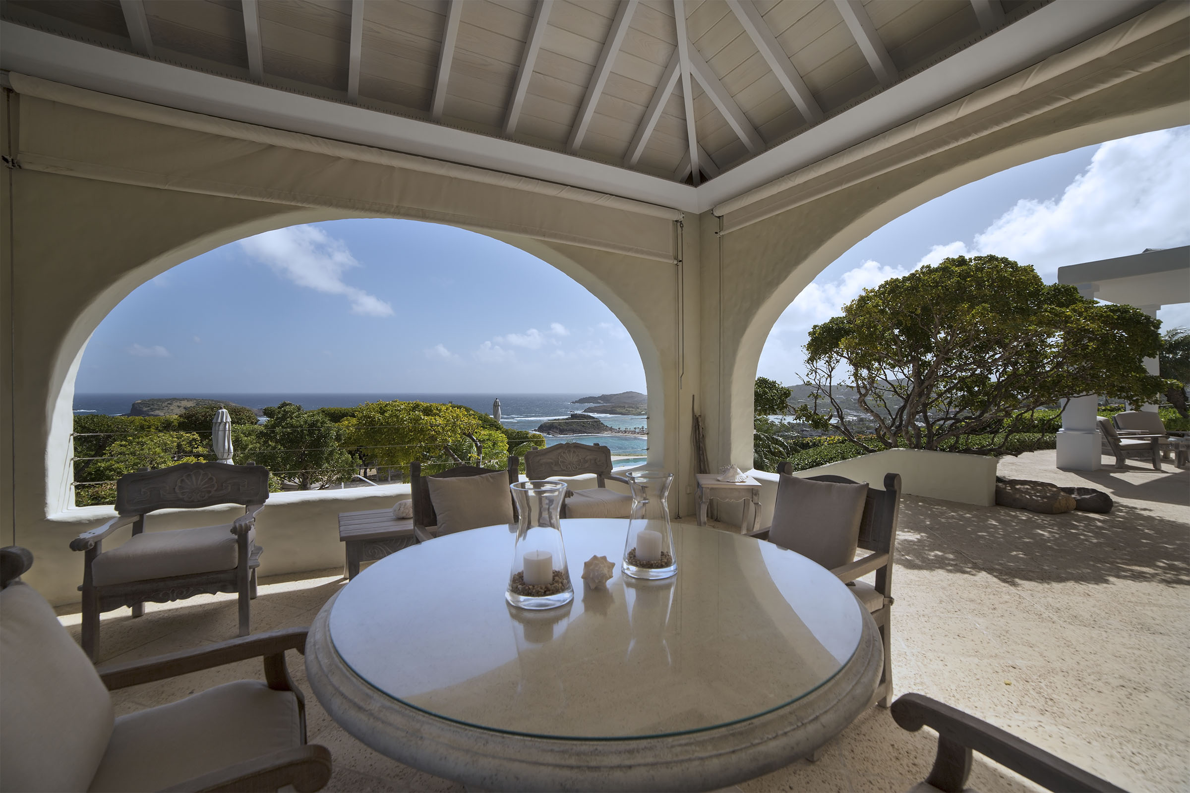 Covered outdoor dining area of a St Barts villa overlooking the turquoise Caribbean Sea, framed by arched openings and soft island light.