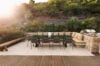 Outdoor dining area with a long table, cushioned seating, and stone walls surrounded by lush greenery at sunset.