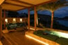 Cozy outdoor lounge with cushioned seating and warm lighting overlooking the ocean and hills at night.
